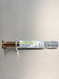 A photo of a short syringe with a brown plunger and a luer-lock cap resting on a brushed metal surface.  The a label is on the glass syringe, it reads Human Papilloma Virus, 9-Valent Vaccine, Recombinant.  Other text informs the reader of the lot number and expiry date.