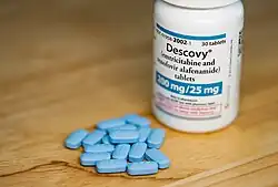 A photo of a small white medicine bottle standing next to a small collection of small sky-blue pills on a wooden table.  The pills have a distinct beveled shape.