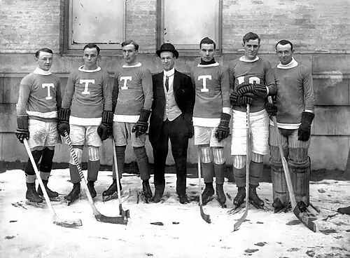 Seven men in row standing in front of brick wall. Six of the seven are wearing hockey sweaters and uniforms