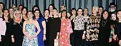 A group of people mostly women stand close to each other and smile at the camera