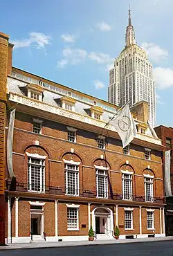 A multi-leveled brick building with many white-paned windows, with a skyscraper visible in the top right corner