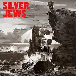 A black-and-white photo of the ocean crashing against a rocky bluff, with multiple stuffed elephants stood on the rocks.