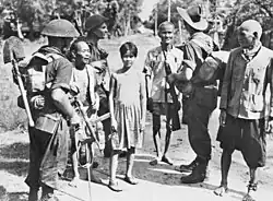 Black and white photo of three men wearing military uniforms and carrying guns with three elderly men and a teenager wearing civilian clothes