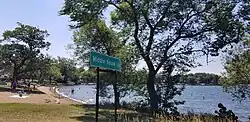 A lake beach surrounded by trees, with people playing in the water in the distance.