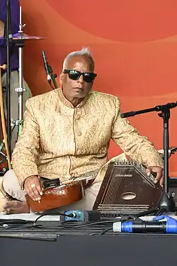Baluni Shrivasta sits on a stage with string instruments resting on his leg and on the floor.