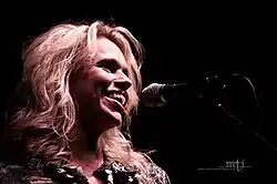 Close-up shot of a woman's face as she sings into a microphone. Her slightly, wavy, blonde hair falls to her shoulders.