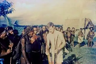 Australian officer casually dressed in only shorts holding a bow and other weapons while others look on