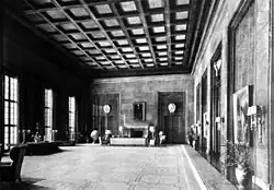 Black-and-white photograph of a large room quite empty. On the back, a fireplace surrounded by five armchairs and a wide couch. A globe is between the leftmost armchair and the left wall. One of three French window lights the globe.