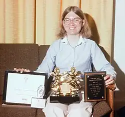 A woman sitting indoors on a couch. She has award statuettes and is holding two award plaques. The first plaque discernibly reads "1984 - Certificate of Merit - River Raid Activision Atari Computers - Best Computer Action Games" and the Electronic Games magazine logo. A matching award statuette next to her leg says "1984 Best Action Videogame River Raid / Activision. Atari 2600." An award statuette on her lap says "Electronic Fun 1983 Hall of Fun". The final plaque reads "Video Game Update - Awards of Excellence - Game of the Year - Atari 2600 - River Raid - By Carol Shaw for Activision - 1983."