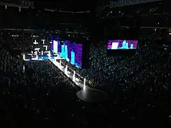 A darkened stage seen from above. Vertical and horizontal white poles of lights rise from both the main stage and from a long catwalk in front of it. Above a large screen above the catwalk shows white-bluish skyscraper images against a purplish night sky