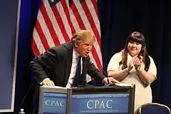 Trump, leaning heavily onto a lectern, with his mouth open mid-speech and a woman clapping next to him