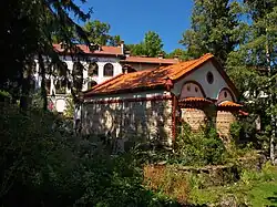 Apse view of a late medieval stone church with a tiled roof and a residential building in the background, all located in a forested area