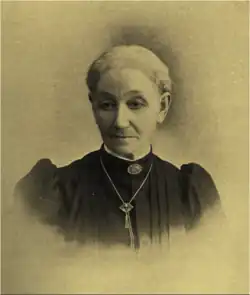 B&W portrait photo of a woman with her white hair in an up-do, wearing a dark blouse, with puffy sleeves and white fringe at the collar, as well as brooch at the neckline and a necklace.