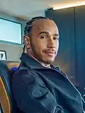 Hamilton is wearing a black zip-top, sitting in a chair, looking and smiling at the camera