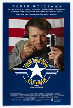 A man wearing military green points with one hand and holds a microphone in the other, against a backdrop of the American flag