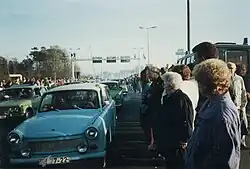 East German Trabant cars driving between dense crowds of people. Metal gantries over the road and a border watchtower are visible in the background.