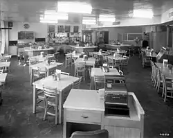 eight or ten light colored wooden tables with chairs seen from behind host station, lit with six bright ceiling lamps