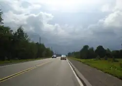 "A rural paved highway running alongside forests and fields. The road is moderately busy. A storm is approaching in the distance."