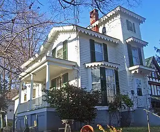 A white house with frilly rooflines lit by the sun from the left, seen from the left and slightly below. It has a tower on the right with a brick chimney.