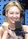 Woman with long, blonde hair wearing large headphones while speaking into a studio microphone. Her hands are lightly clasped in front of her as she smiles brightly, conveying a cheerful and engaging demeanor.