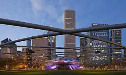 Evening view of a green lawn under a lattice with a lit metal bandshell against a backdrop of skyscrapers