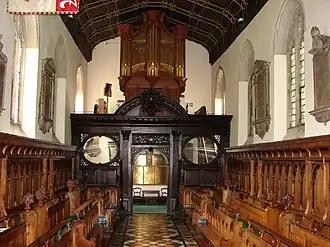 An organ with gilded pipes on top of a decorated wooden screen, with two rows of wooden benches on either side of an aisle