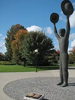 Statue with two arms in the air, each holding a brimmed hat. A bronze plaque on the ground stands in front of the statue