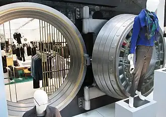 A view of the bank vault door and opening. The circular door has been opened and is hanging to the right of the vault opening. There are clothing displays on the door.