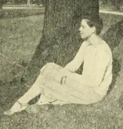 A young white woman seated on grass outdoors, wearing light-colored clothing