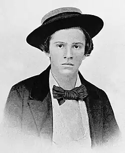A young North American boy of the 19th century. In this black-and-white photograph, the boy looks into the camera nervously, a straw boater perched upon his head. He wears a white shirt, dark jacket and a large, dark-coloured bow tie.