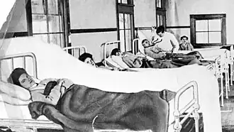 A white woman with dark hair is lying in a hospital bed; she is looking at the camera
