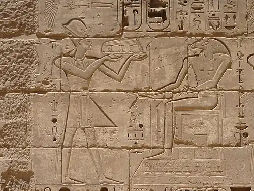 Relief of a man with a crown holding a tray of food in front of a seated man with the head of a ram