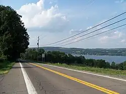 A two-lane highway runs alongside a large lake. The road and the lake are separated by a row of trees and small brush.