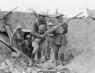 A black and white photograph of a group of men in military uniform outside a dugout in a trench, with one man helping another display a gun for the camera