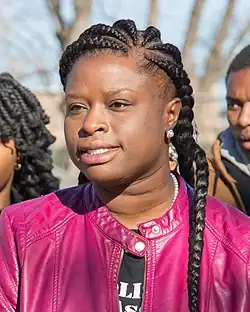 A woman with braided black hair and a purple leather jacket faces TV cameras.