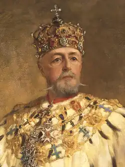An elderly, bearded man in ornate robes, wearing a jewelled crown surmounted by a cross, looks straight out of the picture.