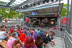 two glassmakers operate kilns and other equipment on a stage, a crowd spectates from seating