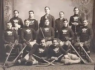 Black and white image twelve men posing for a photo. They are arranged in three rows, five men in the back and middle row, two in the front. The back row is standing, the middle row sitting, and the front row lying on their side towards each other. Ten of the men are wearing a dark wool sweater with an overlapping "P" and "L" with wings, each holding a hockey stick and wearing skates. The other two men are dressed in suits.