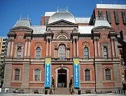 A 3-story, brick and red stone, second empire style building with a slate roof. The building is symmetrical with the entrance center and a blue and yellow banner on either side.