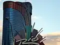 Distant ground-level view of an all-glass tower; the tower has several setbacks near its roof. The glass is mostly a dark blue color, but some windows are tinted red to form a stripe down the side of the building. A prominent "Rio" sign is visible in the foreground.