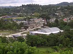 View across a wide, shallow valley with scattered buildings interspersed with dense patches of trees, and open, grassy spaces. The valley rises to a low, forested ridgeline. One of the hills supports a tall broadcasting mast.
