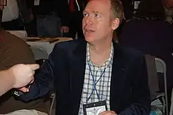 A man with the shot focused on his upper body. He is wearing a dark colored jacket and a white and gray shirt.