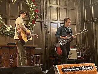 Two people playing guitars, one acoustic and one electric, on a stage. The one on the left is near a microphone but isn’t singing into it