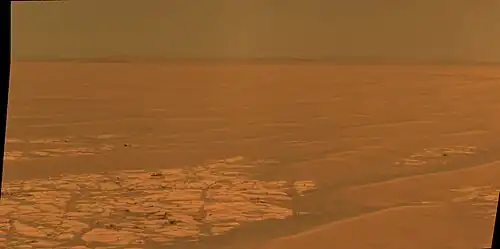 Figure 4. The plain at a field of "plains ripples" soils. Fields of plains ripples cover many square kilometers. The hills on the horizon are the rim of Endeavour Crater. Each plains ripple is a sloped mound of soil, usually around 25 cm tall, 2 m to 4 m wide, and longer than they are wide. The ripples' long axes are roughly aligned north-to-south since the prevailing winds are (like the equatorial trade winds on Earth) from east to west. Each ripple can join with others or be separated by patches of lighter Meridiani sediment outcrop (for example, in the left foreground). A scattering of loose blocks sits on the foreground sediment. Most block are ejecta from meteorite impacts. Plains ripples are smaller than dunes, "armored" by a layer of hematite spherules, and, hence, have exceedingly slow migration rates. Image taken on Sol 2314 (2010-07-28).