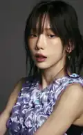 Woman with long, dark hair and soft bangs. She has a smooth, light complexion and is wearing subtle makeup with a glossy lip. Her outfit features a textured, floral-like pattern in shades of lavender and blue.