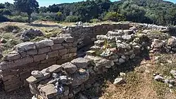 Photograph of the very partial remains of an ancient Greek tomb: a stone passageway in the open air leading to a circular chamber.