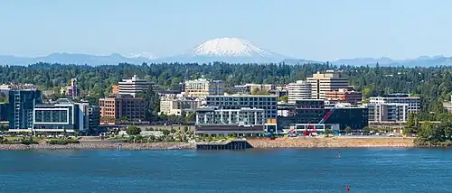 Several mid-rise buildings against the shore of a large river with Mount St. Helens (a snow-capped volcano) in the background behind rows of evergreen trees.
