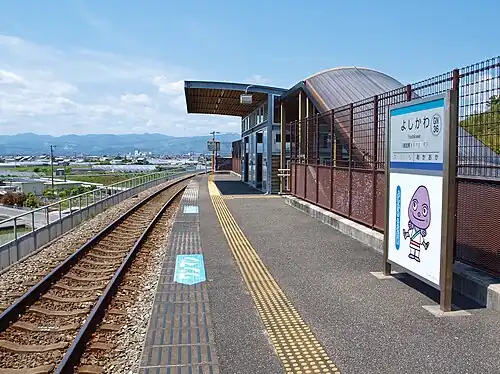 A view of the station platform and track. There is a picture of the station eel mascot character under the station name board.